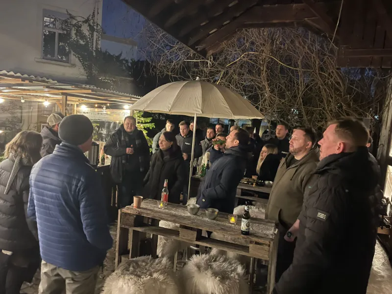 Gruppe in Winterjacken abends an Holztischen unter einem Sonnenschirm