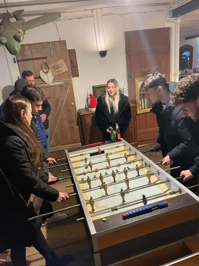 Sieben Personen spielen Tischkicker in einem Raum mit Deckenbalken
