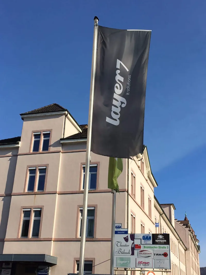 Schwarze layer7-Fahne am Fahnenmast vor einem Altbau bei blauem Himmel