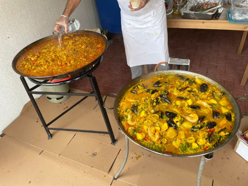 Zwei große Paella-Pfannen im Freien, eine behandschuhte Hand gibt etwas hinein