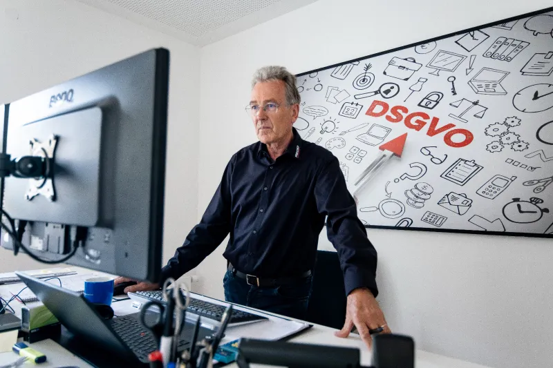 Kollege steht am Schreibtisch, dahinter ein Wandbild mit dem Schriftzug DSGVO