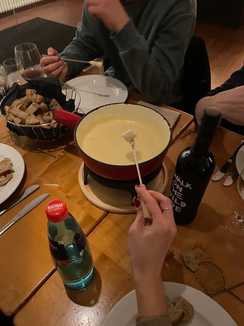 Käsefondue auf einem Holztisch, eine Hand taucht Brot in den Topf